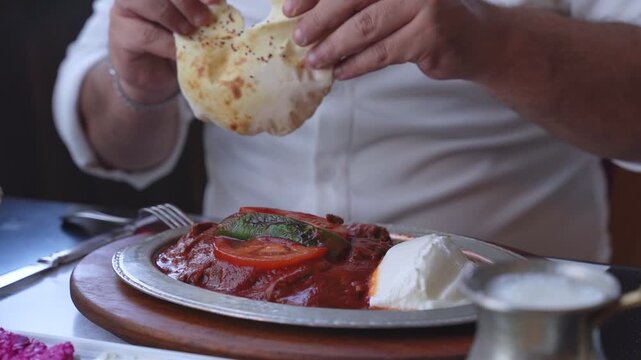 Close up shot of a man in a white shirt tearing fresh flatbread over a traditional Turkish Iskender kebab plate in a restaurant. The dish features meat, tomato sauce, and yogurt under natural light.