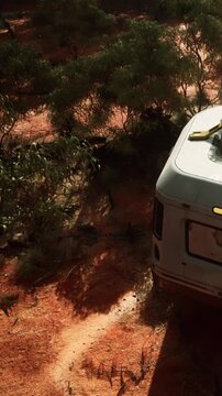 A caravan is parked on a dirt road in a natural area with scattered bushes and trees. The ground is reddish, and sunlight touches the scene. It is a quiet moment in the outdoors.