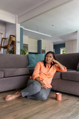 Obraz premium Mature adult female sitting on floor in living room leaning against gray sofa with coral mug
