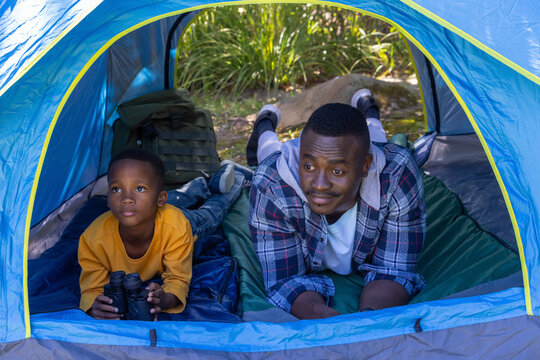 African American father in plaid, son in yellow lying on pads in blue tent holding binoculars