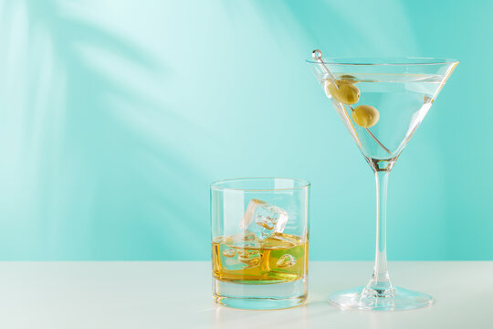 Glass of whiskey with ice and martini with olives on blue background