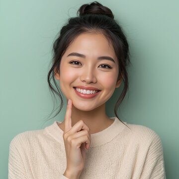 A radiant young Asian woman with a charming dimpled smile touches her cheek while wearing a cream-colored knit sweater against a solid sage green background.