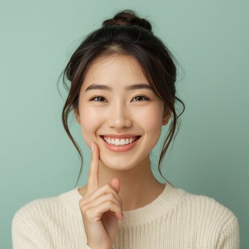 A radiant young Asian woman with a charming dimpled smile touches her cheek while wearing a cream-colored knit sweater against a solid sage green background.