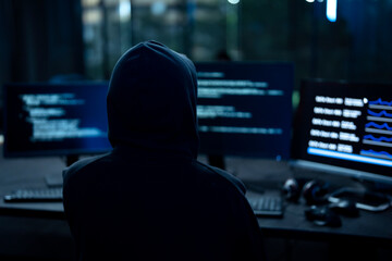 Male computer hacker in black hoodie typing code on computer in a dark room with light reflection...