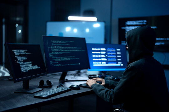 Male computer hacker in black hoodie typing code on computer in a dark room with light reflection on glasses.