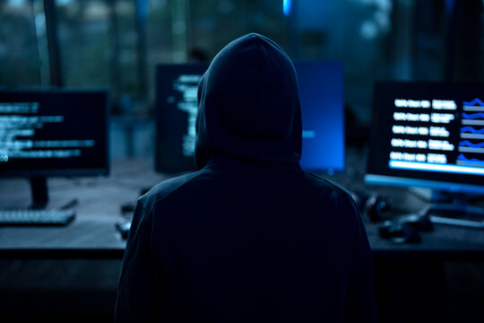Male computer hacker in black hoodie typing code on computer in a dark room with light reflection on glasses.