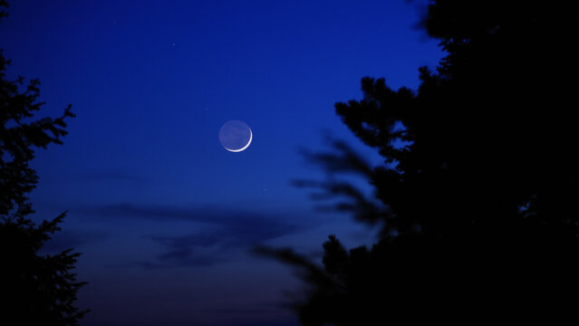 Blue hour time with crescent Moon, stars and planets above landscape silhouettes.