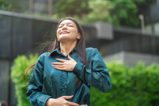 Mental health, happy young asian businesswoman taking a deep breath of fresh air with hand on chest standing outdoor, Relief, relaxing, corporate wellness, mindfulness and success concept