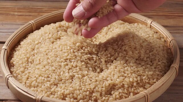 Hand gently touching and pouring unpolished brown rice in a bamboo basket, overhead close-up