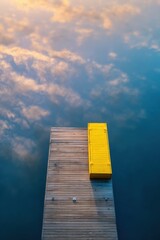 Fototapeta premium Wooden pier with yellow bench on calm lake surface. Aerial view of dock reflecting sky and clouds. Serene nature landscape for vacation, travel meditation and peaceful rest concept.