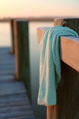 Obraz premium Light blue towel hanging on a wooden dock railing at sunset. Coastal morning scenery with soft sunlight glowing on the fabric. Relaxation, summer vacation and beach lifestyle concept.