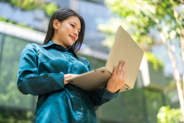 Fototapeta premium Blogger smiling asian woman working on laptop outdoors in green park, Freelance digital nomad woman writing online content, e-learning, Modern remote work internet and work life balance concepts