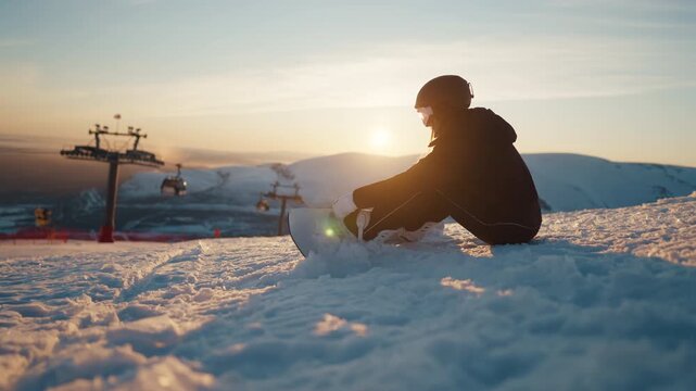 Snowboarding in modern ski resort, sportswoman sitting at top of slope in winter . Rest in mountains, woman snowboarder preparing to ride, modern cable car for rising in background, weekend trip