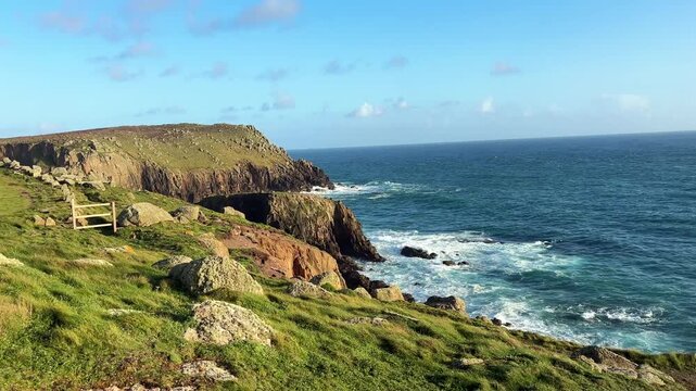 Ground level view of grassy rocky cliffs at Lands End Cornwall with Atlantic waves crashing along rugged shoreline under bright blue sky creating scenic coastal view