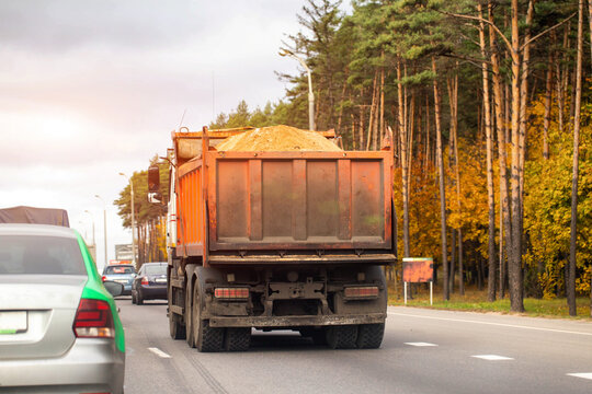 Rear view of orange dump truck carrying sand on highway with other vehicles in traffic, autumn forest lining side of road under overcast sky, transportation of construction materials.