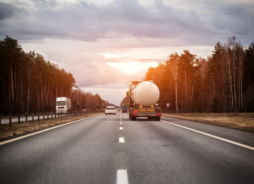 White tanker truck transporting flammable liquid on asphalt highway, surrounded by dense pine and birch forest, under overcast sky, showing essential cargo transportation, evening sunset. Copy space