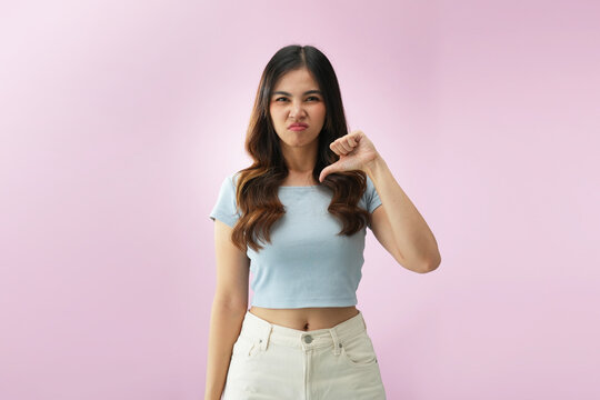Disappointed Indonesian woman showing thumbs down gesture sideways with upset facial expression on pink background, illustrating negative opinion, dissatisfaction, and rejection concept.