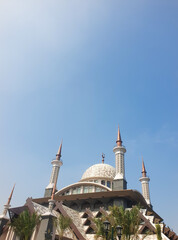 A striking view of a modern mosque featuring a patterned central dome, sharp spires, and traditional crescent symbols against a blue sky. © YB14