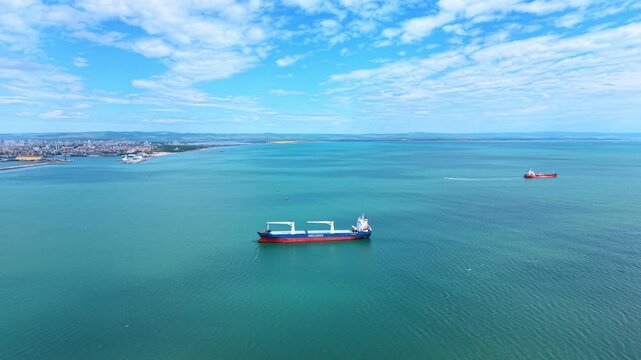Aerial view of cargo bulk carrier ship in turquoise sea. High angle drone shot of a large blue and red bulk carrier vessel anchored near a coastal port city under a cloudy sky.
