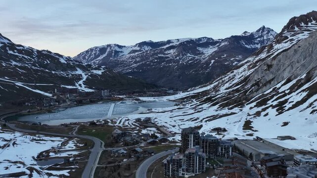 Evening drone view of Tignes 2100 in the French Alps, showing illuminated mountain resort buildings and hotels surrounded by dramatic alpine peaks under twilight sky atmosphere