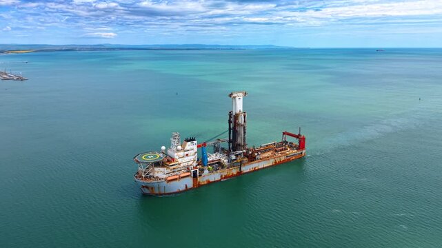 Industrial offshore drillship navigating calm turquoise sea water under clouds. Aerial view of a large drilling vessel with central tower and helipad sailing in open ocean towards the horizon.