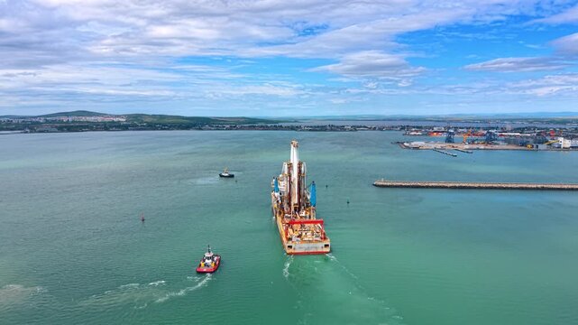 Aerial view of offshore drilling vessel escorted by tugboat. Drone shot of industrial drillship with derrick tower and tugboat on turquoise sea near commercial port background.