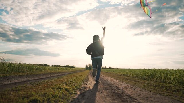 Little girl running along grassy dirt field. Child flies colorful kite. Backlit girl enjoys running safely outdoors. Dark silhouette reveals freedom. Wind lifts kite over beautiful summer field.