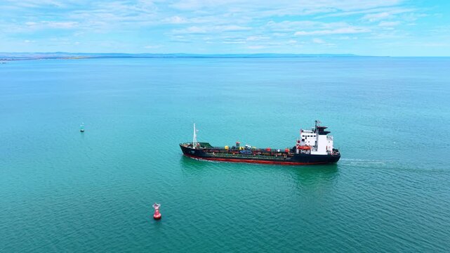 Aerial view of industrial tanker ship sailing in turquoise ocean. Drone view of industrial tanker ship navigating blue sea with red and green navigation buoys under cloudy sky.