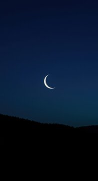 A dramatic deep blue sky with a sliver of bright crescent moon hanging over a dark, silhouetted landscape at midnight ,night ,mystery ,waxing