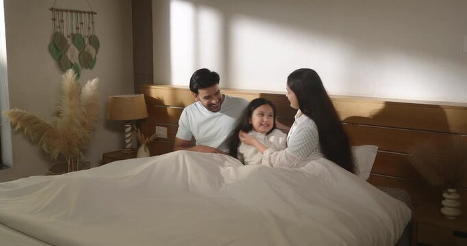 Indian man woman sitting on cozy bed do tickling to girl talk speak show love care enjoy day living room home. Young adult mom dad male female play kid child spend family time have fun joy house place