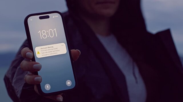 Woman Holding Smartphone With Hurricane Warning Notification Near Stormy Coastline at Dusk, Emergency Alert and Natural Disaster Preparedness
