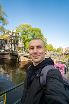 Young tourist man smiling, taking a selfie while visiting amsterdam city, exploring canals and bridges