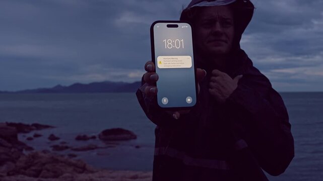 Woman Holding Smartphone With Hurricane Warning Notification Near Dark Ocean Shoreline at Dusk, Severe Weather Risk and Emergency Preparedness