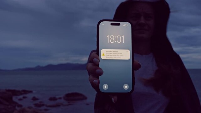 Smartphone Hurricane Warning Notification Held by Woman Near Dark Coastal Landscape, Severe Weather Forecast and Disaster Preparedness