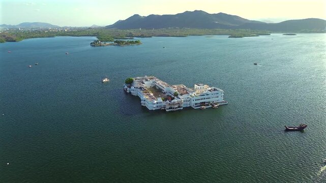 A high-angle wide static shot of the iconic white marble Taj Lake Palace floating on the calm waters of Lake Pichola, surrounded by hills and boats in Udaipur, Rajasthan, India