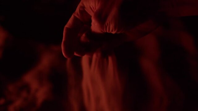 Close up of a hand pouring fine sand through fingers at night, illuminated by a warm red fire light. Concept of time passing, mortality, or mystery in a dark, atmospheric environment