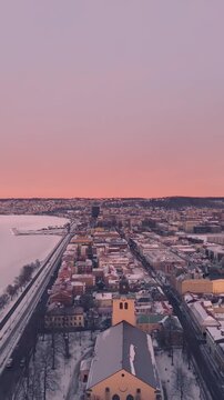 Stunning vertical aerial footage over snowy J&ouml;nk&ouml;ping, Sweden, reveals Sofia Church and frozen V&auml;ttern Lake below a pink-orange sunset sky. Ideal for mobile, stories, and social platforms.