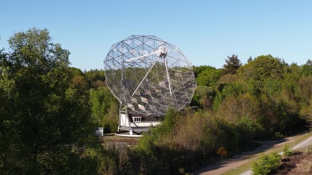 Large dwingeloo radio telescope rotating slowly while searching for astronomical signals from deep space, surrounded by lush green forest under a clear blue sky on a sunny day