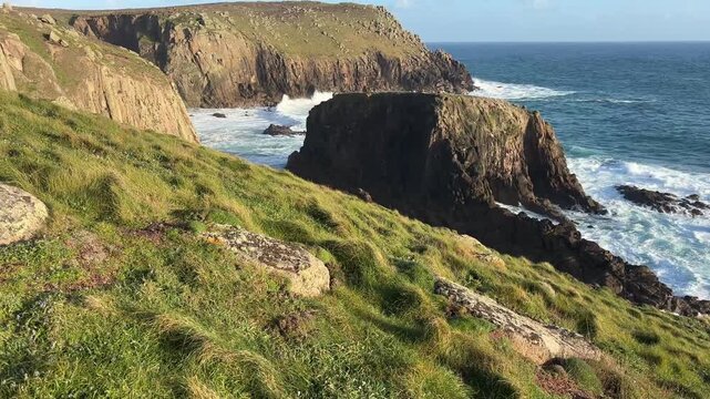 Coastal view of grass covered cliffs and rocky outcrops at Lands End Cornwall with waves crashing against shore under clear sky creating scenic natural coastline