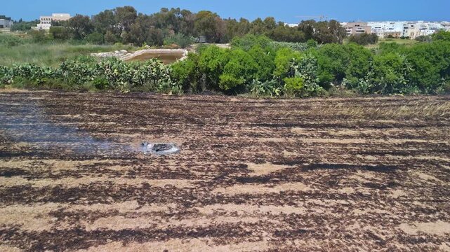 In Attard, Malta, a burnt field with charred ground and lingering smoke lies near suburban buildings and surrounding vegetation in a drone view after a controlled burn.