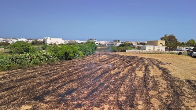 In Attard, Malta, a firefighter works to extinguish a field fire on scorched ground amid smoke and dry vegetation during a hot daytime emergency, captured from a drone perspective.