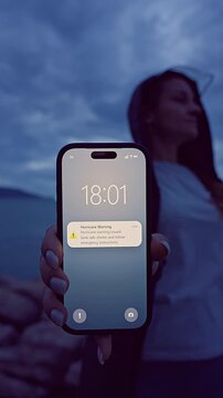 Vertical Shot of Woman Holding Smartphone With Hurricane Warning Near Stormy Coastline at Blue Hour, Severe Weather Awareness and Emergency Safety