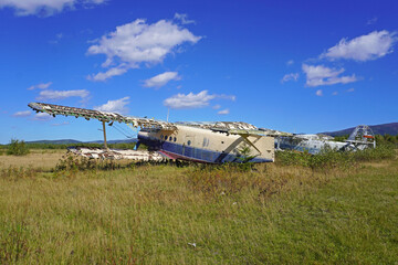 Fototapeta premium Abandoned airplane in a grassy field. Old aircraft fuselage with broken wings stands in tall grass under blue sky with clouds, hills in background, no people.