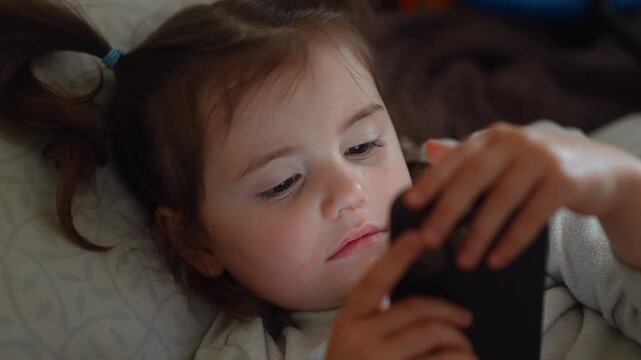 A girl lies on a bed, focused on her smartphone. The girl seems engaged with the device, as her fingers manipulate the screen.