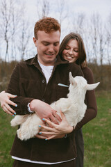 Fototapeta premium Smiling couple outdoors cuddling a white goat kid in a grassy field, warm interaction, soft natural light, rustic countryside mood, joyful companionship.
