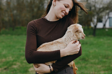 Fototapeta premium Joyful woman outdoors holds a young goat kid in her arms, smiling warmly as they share a tender moment in a green park, showcasing countryside lifestyle and a gentle bond.
