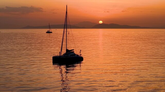 A silhouette of a catamaran sailboat on calm ocean during a golden sunset in the mediterranean sea, Greece, as a background