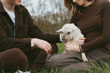 Fototapeta premium Lifestyle outdoor scene shows a couple sitting on grass, gently holding a goat kid, a calm interaction with animal and nature, warm smiles, casual clothing, peaceful family vibe and connection