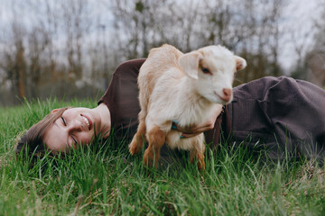 Fototapeta premium A woman lies on green grass beside a playful goat kid in a rural field, enjoying a calm moment outdoors. A simple scene of companionship, nature, and relaxed leisure, highlighting rural lifestyle and