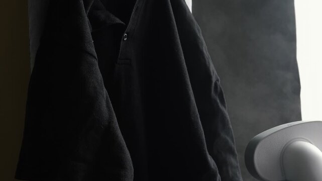 A garment steamer removes wrinkles from a black polo shirt. White steam effectively cares for the fabric at home.